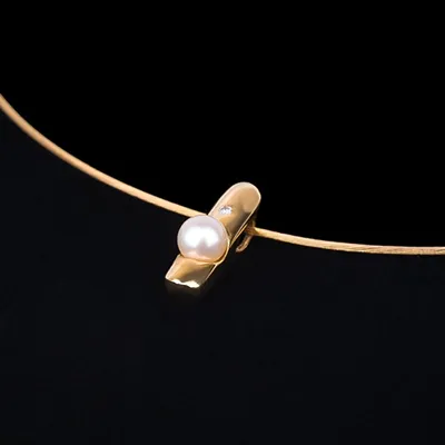 Produktfotografie Schmuck – Gold auf Schwarz Eine Nahaufnahme eines glänzenden goldenen Schmuckstücks mit einer weißen Perle und einem kleinen Diamanten, perfekt ausgeleuchtet auf einem kontrastreichen schwarzen Hintergrund. Die Schärfe und Detailgenauigkeit heben die Materialität hervor.